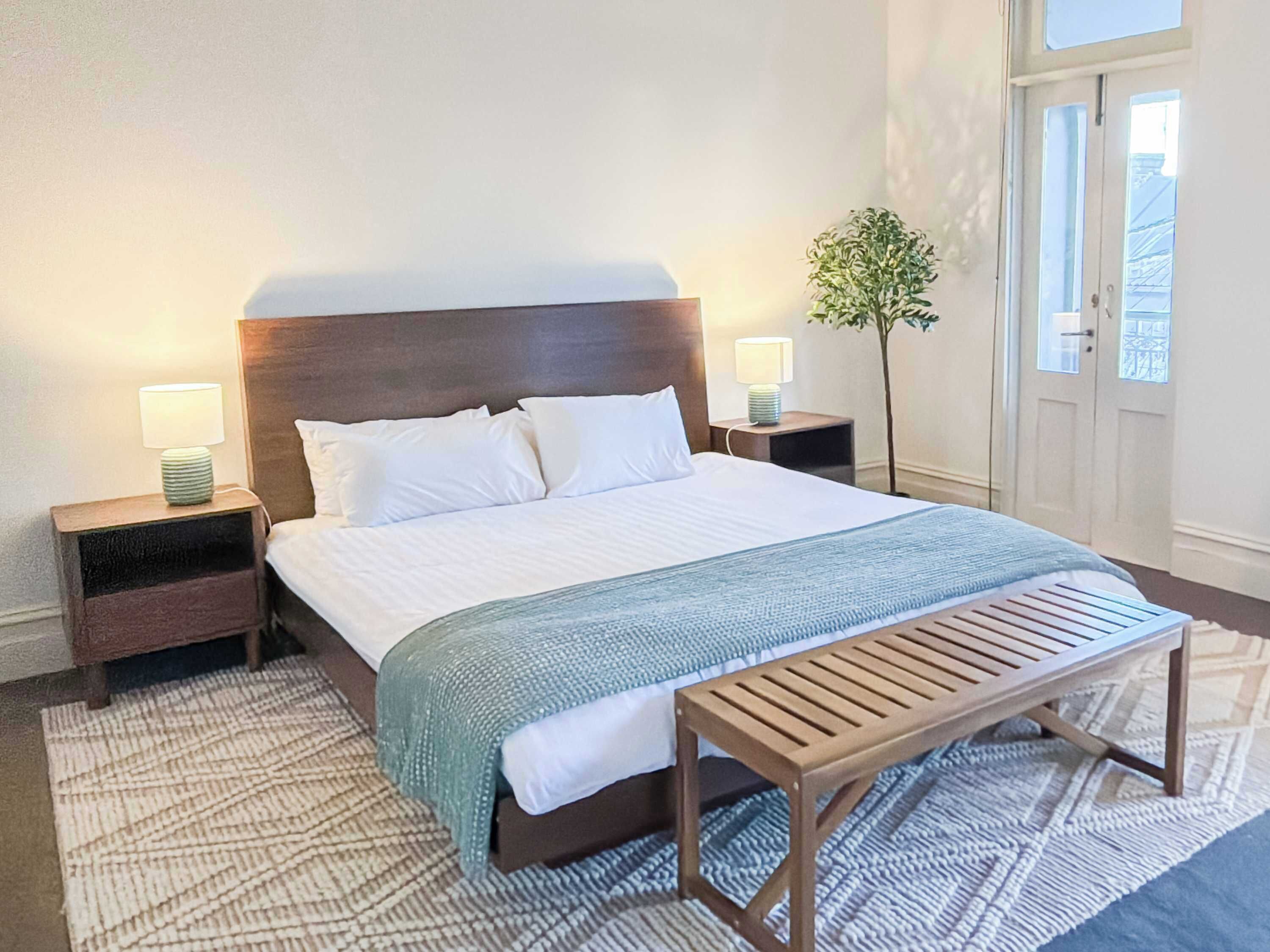 A neatly made bed with white linens, two bedside tables with lamps, a bench, and a potted plant.