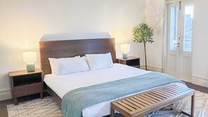 Una cama bien hecha con sábanas blancas, dos mesitas de noche con lámparas, un banco y una planta en maceta.