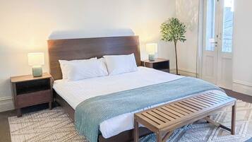 A neatly made bed with white linens, two bedside tables with lamps, a bench, and a potted plant.