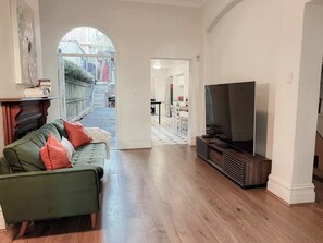 A living room with a green sofa, a flat-screen TV, wooden flooring, and a view through an arched doorway to a backyard. - Huge Heritage Terrace, Walk to KX, Parking, BBQ (Darlinghurst)