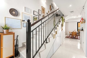 A staircase with a black railing, a dining area with a table and chairs, and a wall decorated with framed pictures and a woven basket.