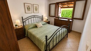 A bedroom with a metal bed frame, a wooden dresser, a nightstand with a lamp, and a window with a view of greenery. - Mandy and Dolly's house with sea view and pool (tertenia)