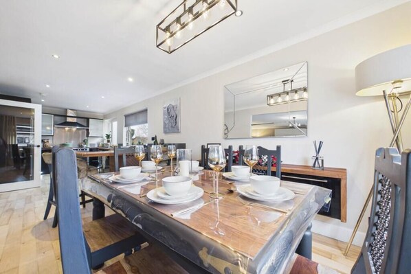 A modern dining area with a long table set for six, a kitchen in the background, and a large mirror on the wall.
