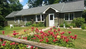 A two-story house with a front porch, a small garden with red flowers, and a wooden fence.