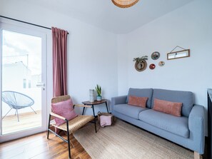 Un salon avec un canapé, un fauteuil, une petite table sur laquelle est posée une plante, et une fenêtre avec des rideaux.