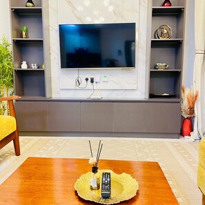 Ein Wohnzimmer mit flachbildfernseher an der Wand, einem Holztisch mit Fernbedienung und einer Flasche, sowie einem Sofa mit gelbem Kissen.