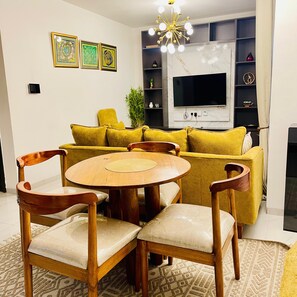 Ein Wohnzimmer mit einer Couch, zwei Holzstühlen, einem runden Tisch, einem Fernseher und einer dekorativen Deckenleuchte.
