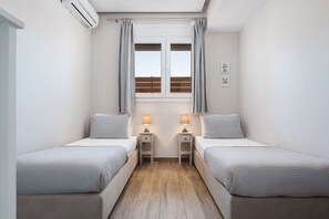 Ein Schlafzimmer mit zwei Einzelbetten, einem fensterbelaubten Fenster, einem Deckenventilator und einem kleinen Nachttisch mit Lampe.