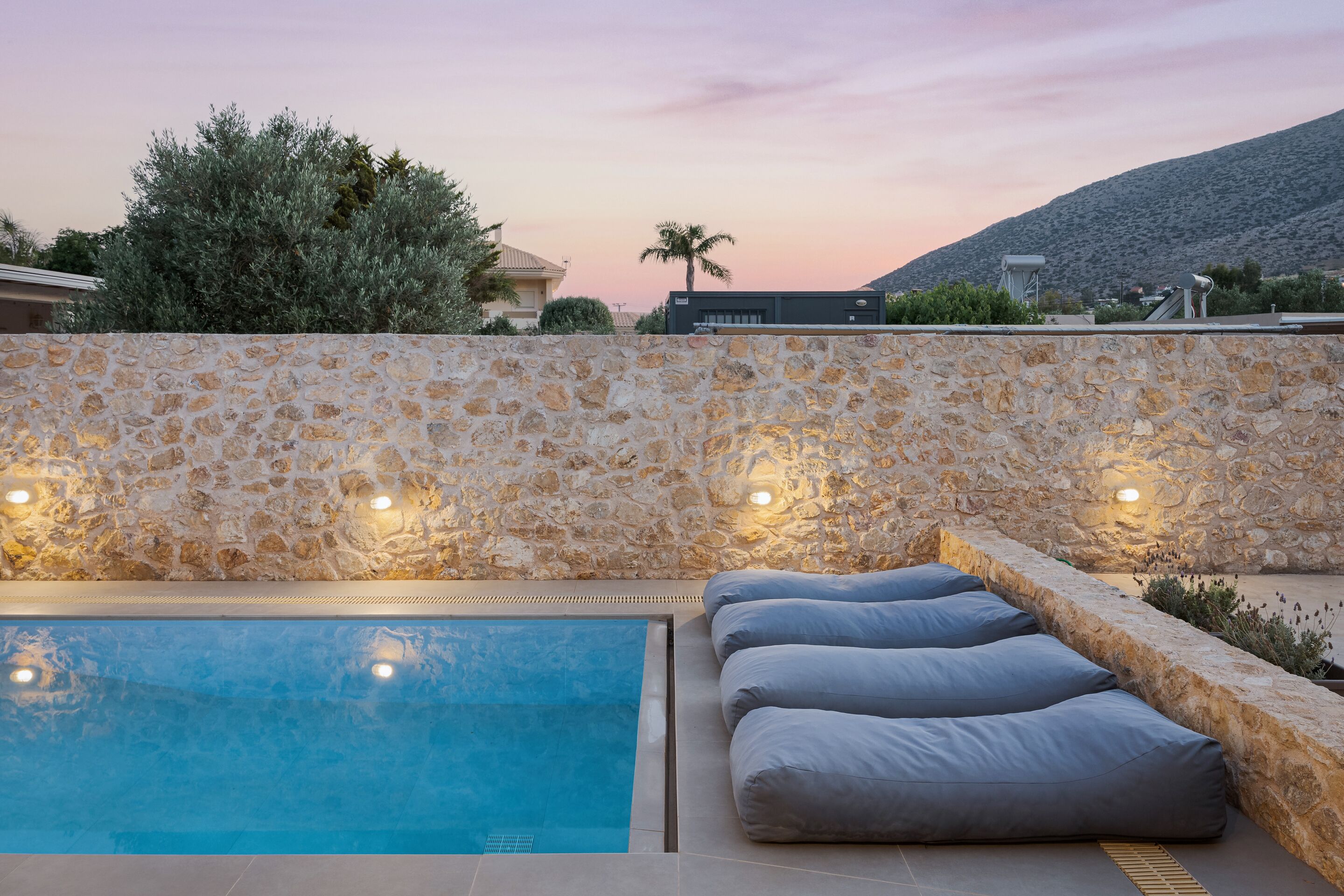 A swimming pool with stone walls and seating, surrounded by greenery and a mountain in the background.