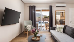 Ein Wohnzimmer mit Flachbildfernseher, einer Couch, einem Sessel, einem Couchtisch mit Blumenvase und Blick auf Pool und Berge.