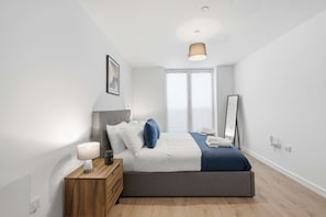 Ein modernes Schlafzimmer mit Bett, Nachttisch, Lampe und einem großen Fenster.