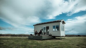 Une petite maison à un seul étage, dotée d’un toit plat et d’une porte d’entrée, située sur un champ verdoyant sous un ciel dégagé.