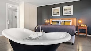 A modern bedroom with a large bathtub, a bed with a wooden headboard, two framed pictures on the wall, and a bedside table with a lamp.