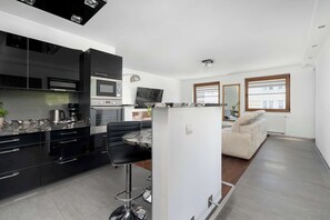 Eine moderne Küche mit schwarzen Schränken, ein Essbereich mit einem runden Tisch und Barhocker, ein weißes Sofa und ein an der Wand montierter Fernseher.