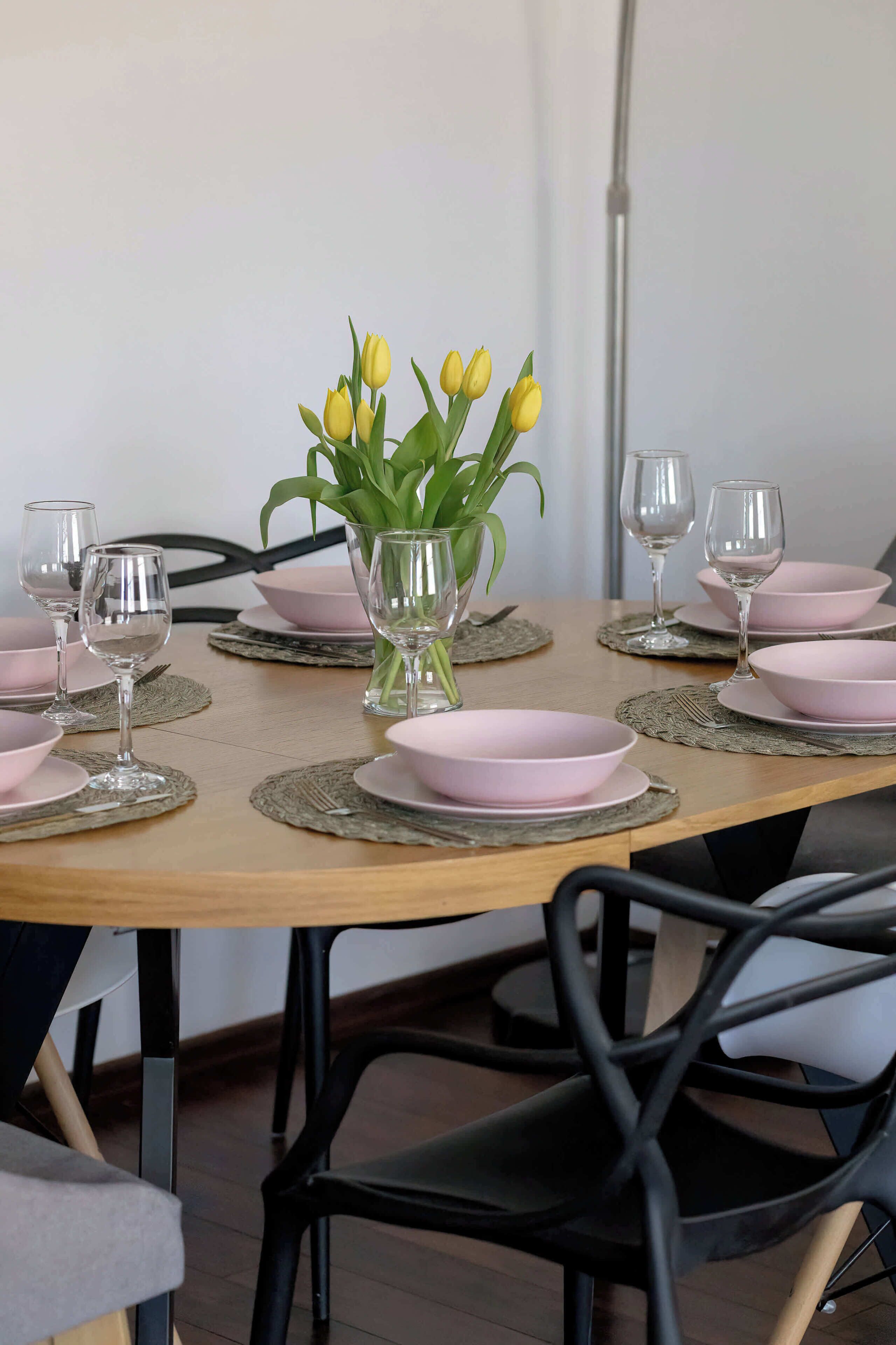 Ein Esstisch, gedeckt mit rosafarbenen Schalen, Weingläsern und einer Vase mit gelben Blumen.