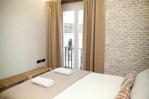 A hotel room with a bed, a bench, a balcony with a view of buildings, and a brick wall. - Homey Seville Brand New Arfe II Apartment Near Cathedral 8 people (Sevilla)