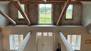 Een loft-achtige kamer met zichtbare houten balken, een visgraatvloer en uitzicht op een groen landschap door twee ramen.