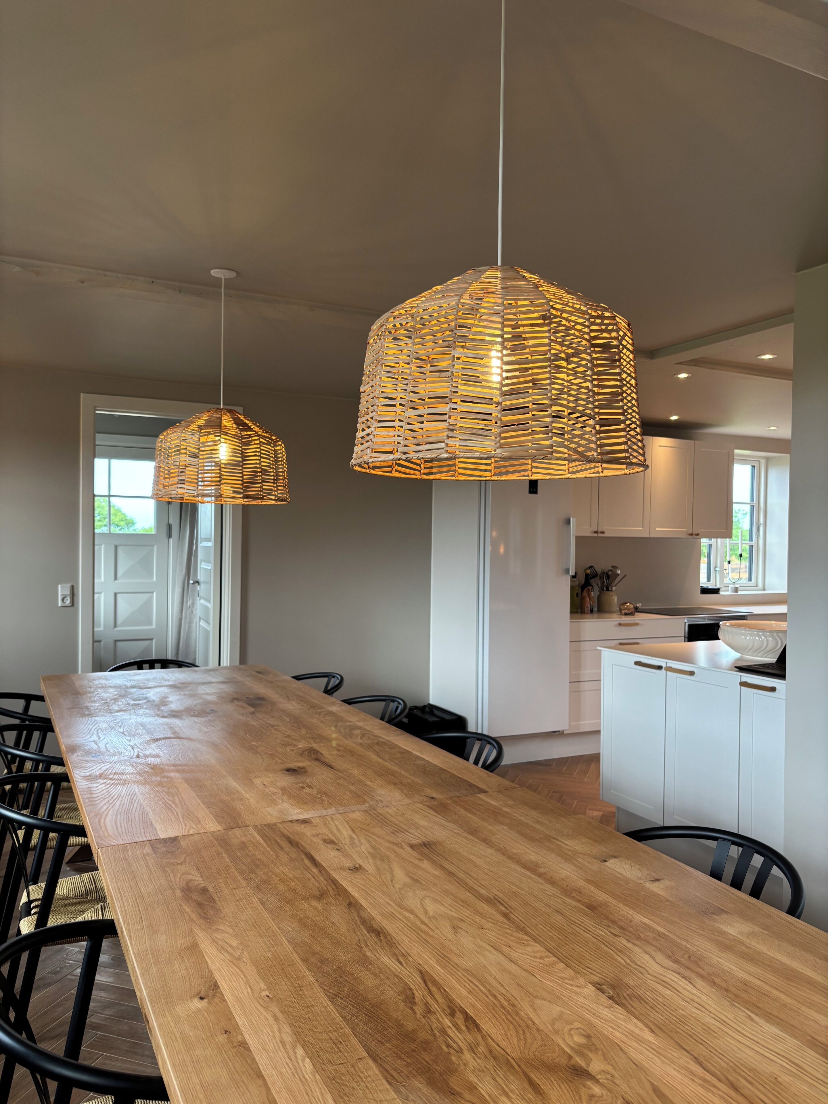 Een moderne keuken met een houten eettafel, twee hangende rieten hanglampen en witte keukenkastjes.