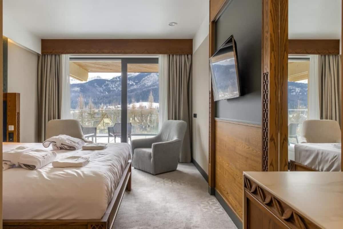 Une chambre d’hôtel avec un lit, deux fauteuils, une télévision et une vue sur les montagnes.