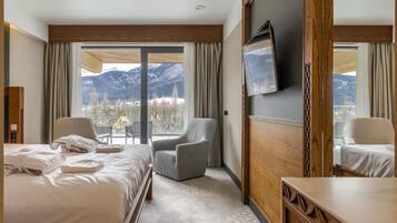 A hotel room with a bed, two armchairs, a TV, and a view of mountains.