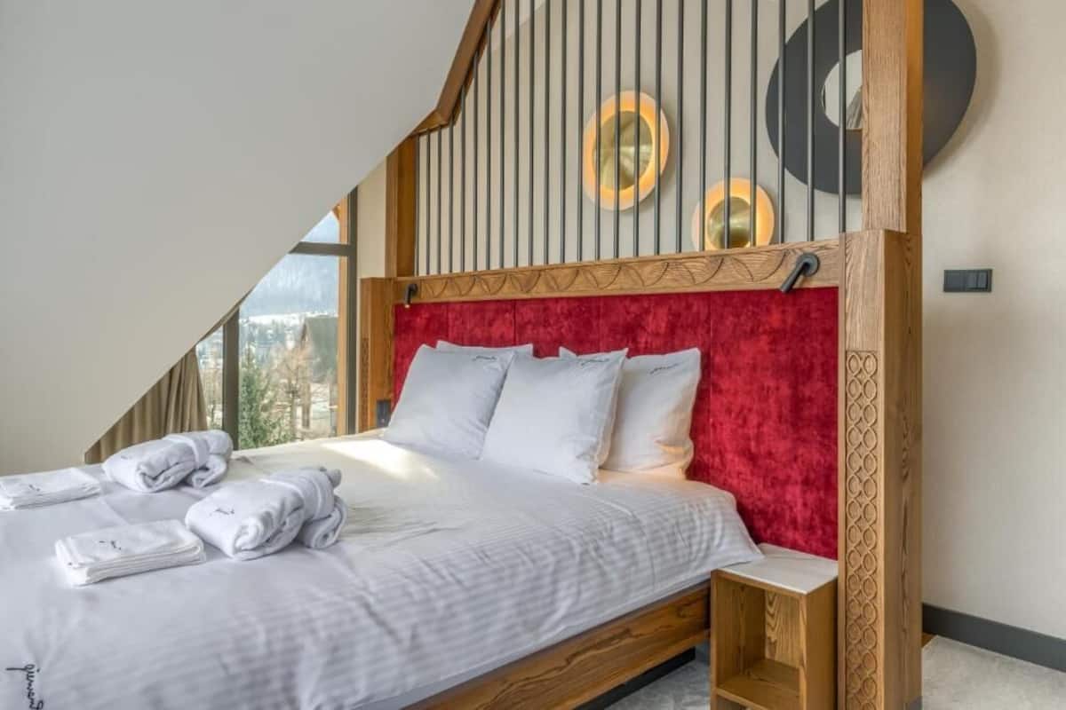 Une chambre moderne avec un plafond en pente, une tête de lit rouge rembourrée, du linge de lit blanc et une vue sur l’extérieur.