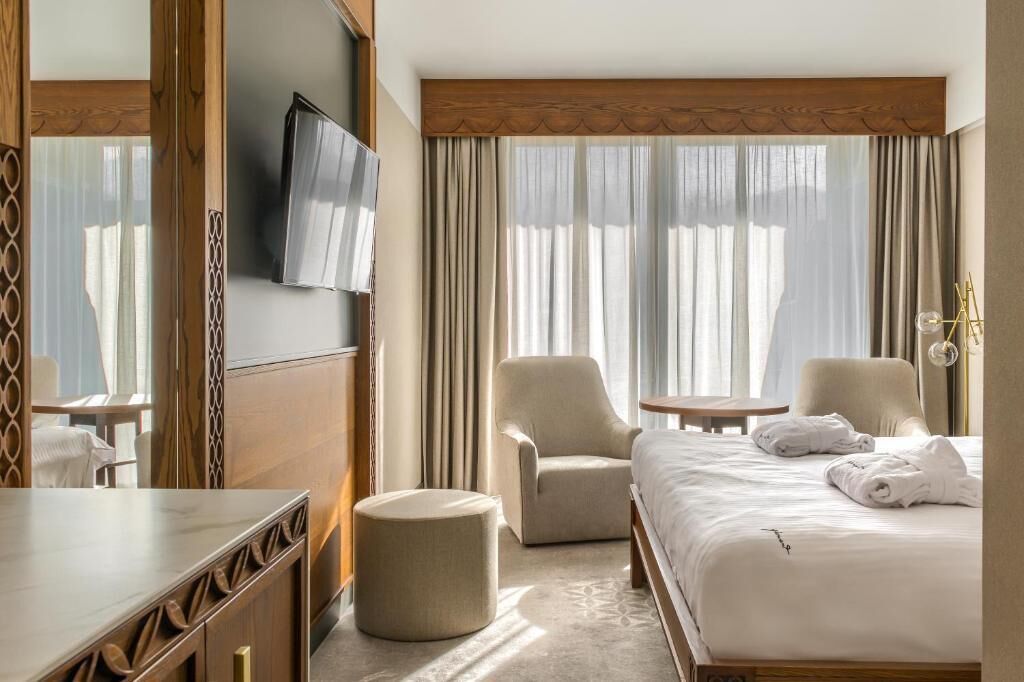 Camera d'albergo con un letto, due poltrone, un poggiapiedi rotondo, una televisione e un mobile in legno.