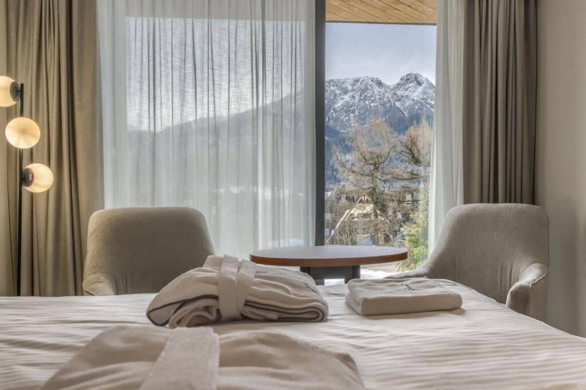 Une chambre d’hôtel avec un lit, deux chaises, une petite table et une vue sur les montagnes à travers de fines rideaux.