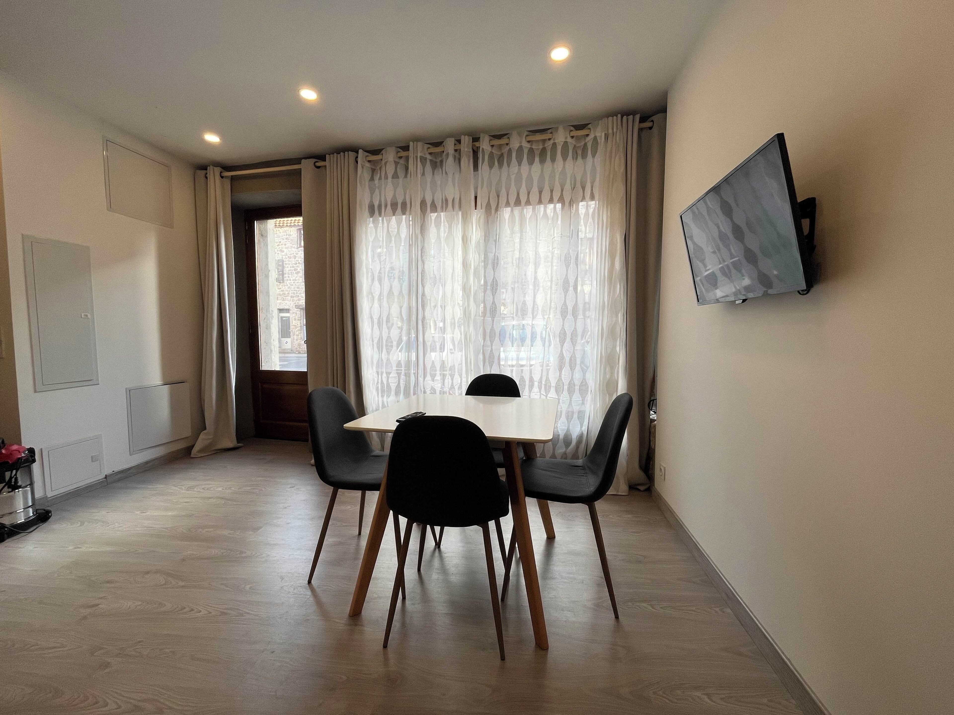 A room with a dining table, chairs, a TV mounted on the wall, and a window with curtains.