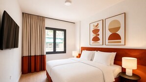 Habitació d'hotel amb un llit, dues làmpades de nit, un televisor, una finestra amb cortines i dues obres d'art abstractes penjades a la paret.