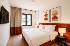Ein Hotelzimmer mit Bett, zwei Nachttischlampen, einem Fernseher, einem Fenster mit Vorhängen sowie zwei abstrakten Wandbildern.