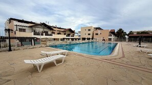 Une piscine entourée de chaises longues, une terrasse en pierre et, au loin, des immeubles à plusieurs étages.