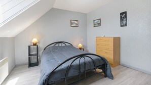 A bedroom with a sloped ceiling, a bed, a wooden dresser, two wall-mounted lamps, and two framed pictures. - Gîte Jersey – Your Vacation Close to the Sea (Bréhal)