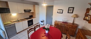 A compact kitchen with a microwave, oven, and dishwasher, a dining table with chairs, and a wooden cabinet with a lamp.