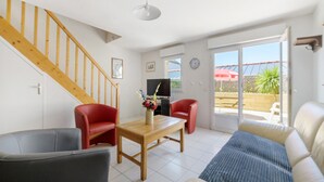 A living room with a wooden staircase, a dining table with chairs, a sofa, and a TV. - Gîte Jersey – Your Vacation Close to the Sea (Bréhal)