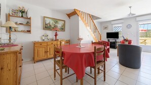 A dining area with a round table, chairs, a wooden cabinet, a TV, and a staircase. - Gîte Jersey – Your Vacation Close to the Sea (Bréhal)