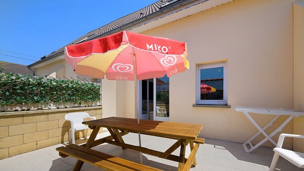 A wooden picnic table with benches, a red and yellow umbrella, and a white plastic chair. - Gîte Jersey – Your Vacation Close to the Sea (Bréhal)