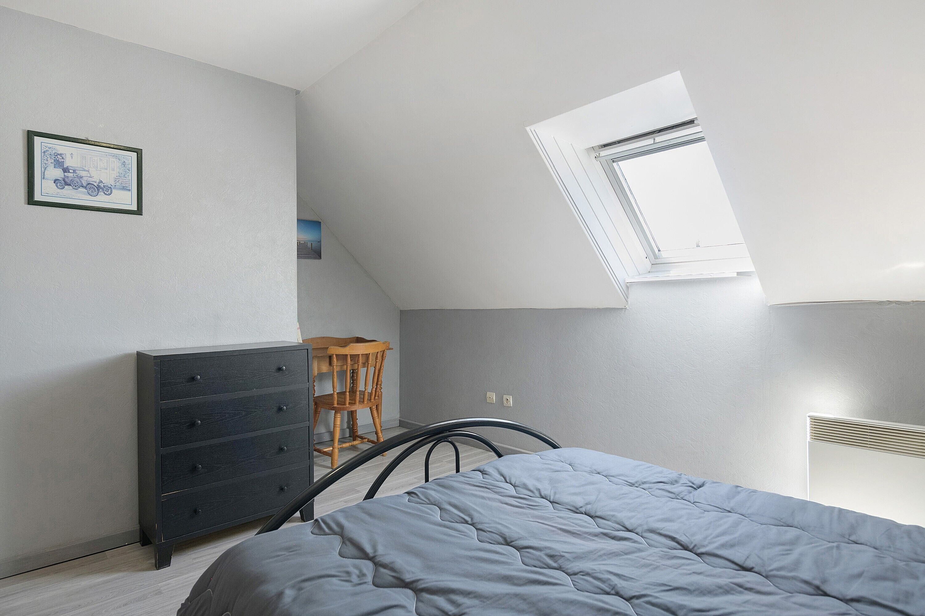A bedroom with a sloped ceiling, a bed, a dresser, a chair, and a radiator.