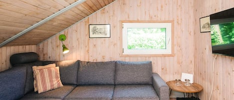 Ein gemütliches Zimmer mit einem grauen Ecksofa, einem runden Holztisch, einem gerahmten Bild und einem an der Wand montierten Fernseher.
