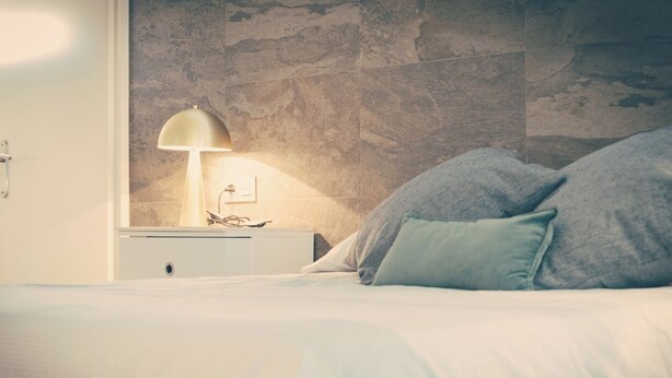 A bedroom with a bed, a nightstand with a lamp, and a textured wall.