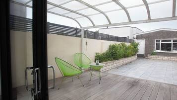 A covered patio area with a wooden deck, two green chairs, a small table, and a glass door leading inside.