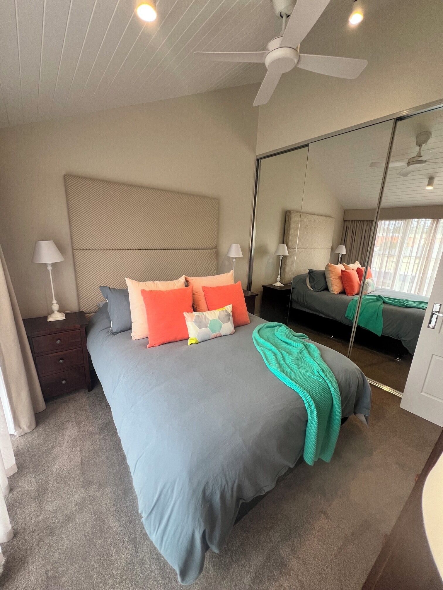 A bedroom with a bed, bedside tables, a ceiling fan, a large mirror, and a view of the outside.