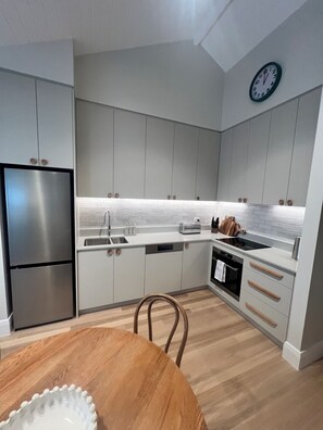 A modern kitchen with stainless steel appliances, white cabinetry, and a wooden dining table. - Seascape Cottage (Glenelg)