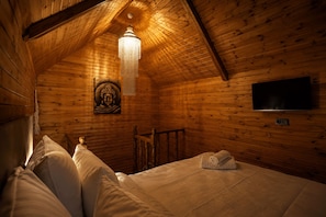 Una habitación acogedora de madera con una cama, una silla de madera, un aplique colgante y un televisor instalado en la pared.