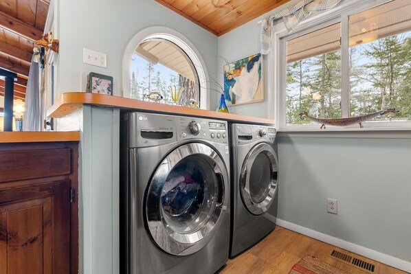 A laundry area with two front-loading washers, a wooden countertop, a window with a view of trees, and a painting on the wall.