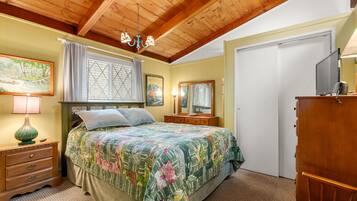 A bedroom with a wooden ceiling, a bed with a floral bedspread, a nightstand with a lamp, a dresser, and a mirror.