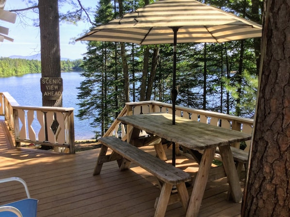 A wooden deck with a picnic table and umbrella, overlooking a lake with trees and a sign reading 'SCENIC VIEW AHEAD'.