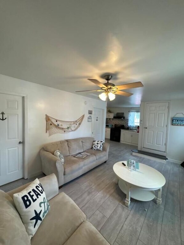 A living room with a sofa, a coffee table, a ceiling fan, and a kitchen in the background.