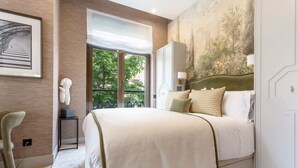 A bedroom with a large bed, a side table, a chair, a painting, and a view of greenery outside. - Exclusive Studio in Salamanca Isla de la Rua I (Salamanca)