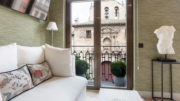 Una habitación de hotel moderna con sofá, televisión de pantalla plana, balcón con plantas en macetas y vistas a edificios históricos.