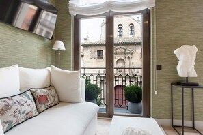 Una habitación de hotel moderna con sofá, televisión de pantalla plana, balcón con plantas en macetas y vistas a edificios históricos.
